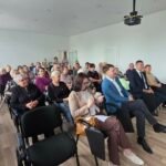 Šviesioje salėje keliomis kėdžių eilėmis sėdi jubiliejinio susirinkimo dalyviai ir svečiai. Jie žiūri į salės priekį; kairėje matyti dideli langai, palubėje – projektorius.