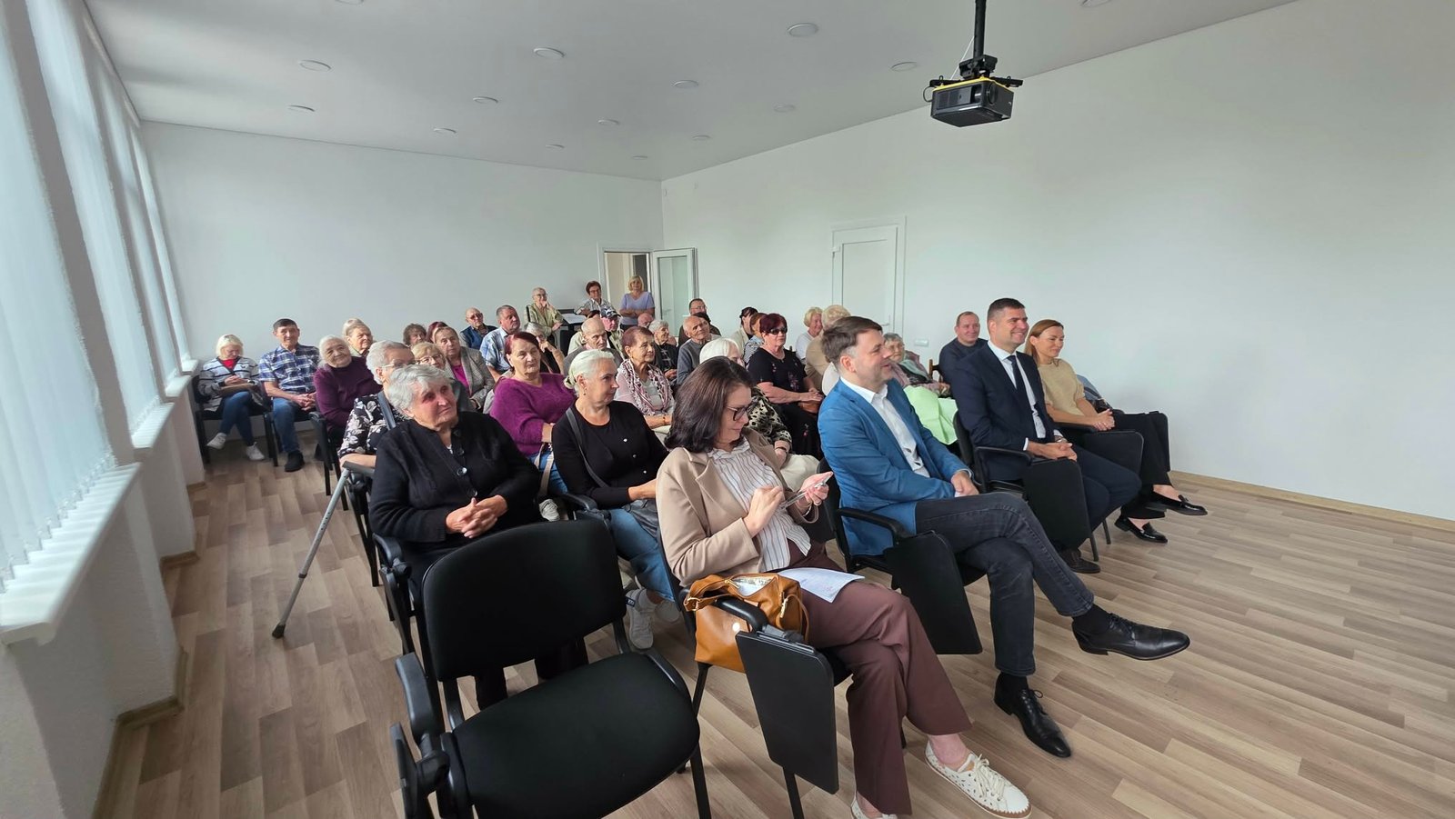 Šviesioje salėje keliomis kėdžių eilėmis sėdi jubiliejinio susirinkimo dalyviai ir svečiai. Jie žiūri į salės priekį; kairėje matyti dideli langai, palubėje – projektorius.